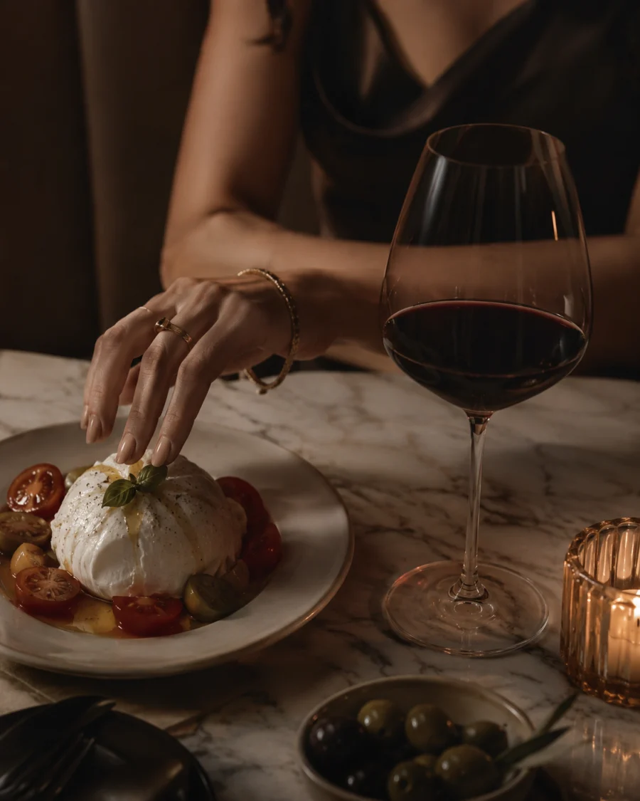 Burrata with tomatoes and basil beside red wine, olives, candlelight, and a guest reaching in